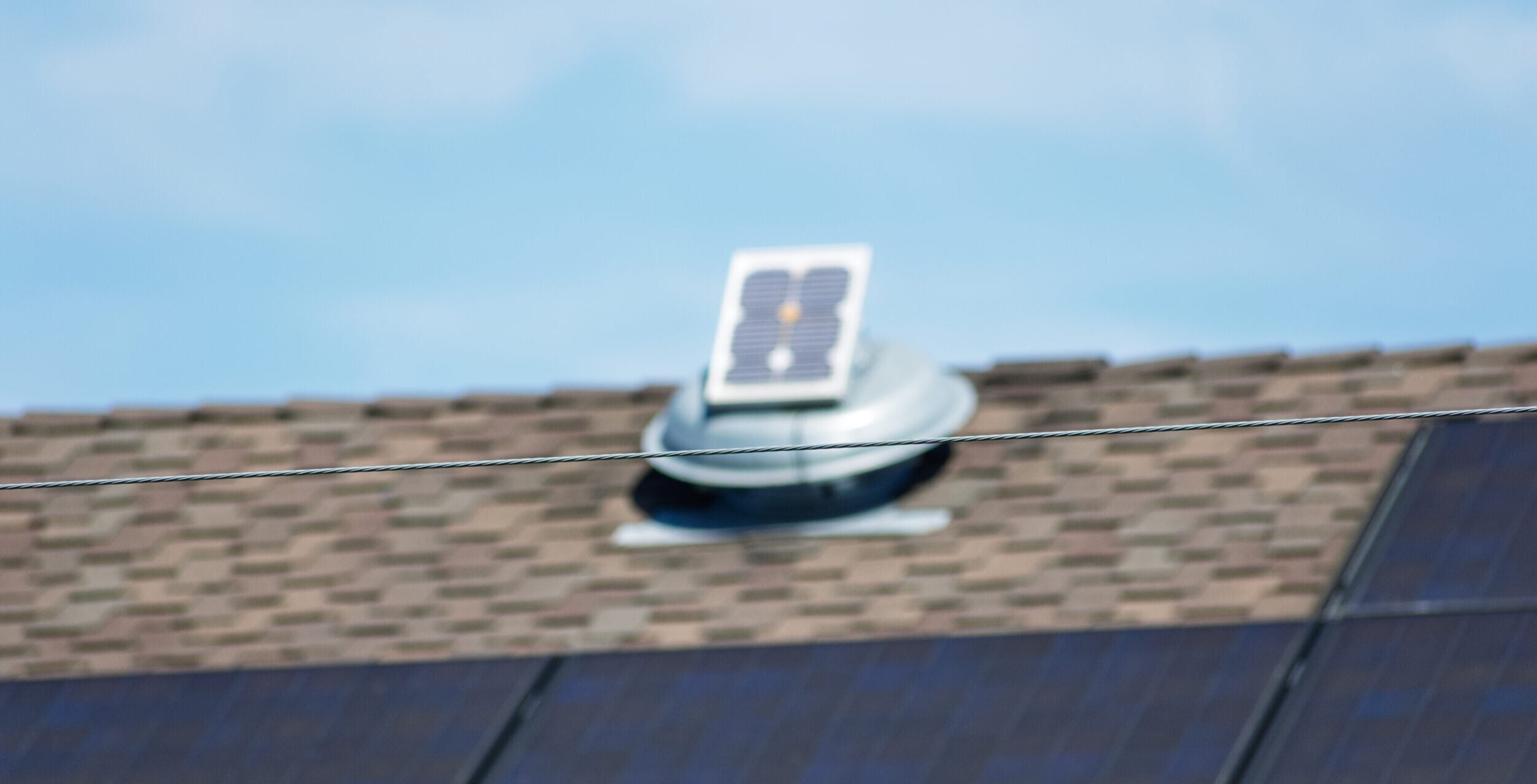 Blurred view of rooftop solar panels and attic fan with sharp focus on foreground power lines under clear blue summer sky.