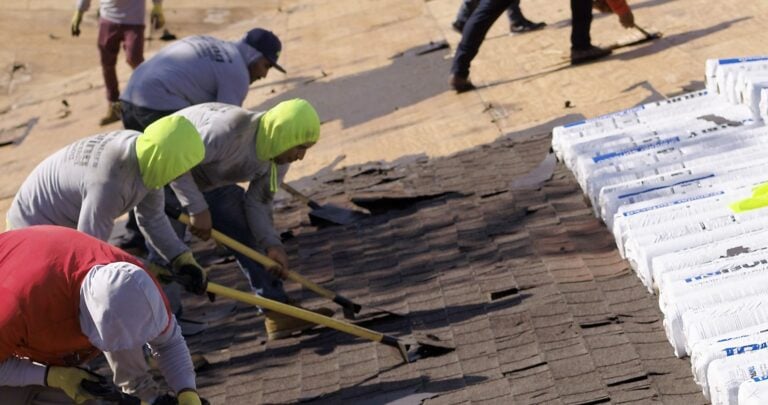 Several workers wearing hats and protective clothing remove old shingles from a roof with tools, while new roofing materials are stacked nearby on the wooden surface.
