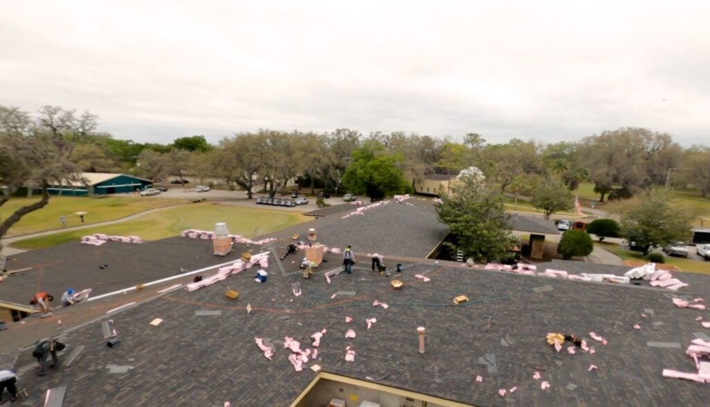 A group of workers installs roofing materials on a large, shingled roof as part of a pitched roof project. Pink insulation is scattered around, and trees and buildings surround the area on a cloudy day.