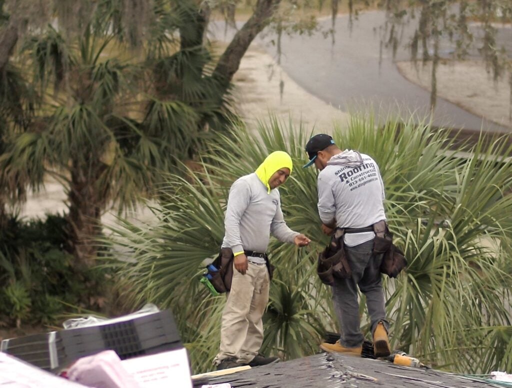 Two construction workers wearing safety harnesses stand on a pitched roof surrounded by palm trees. One wears a bright yellow hood, and both are focused on a task, with roofing materials for a flat roof replacement visible nearby.