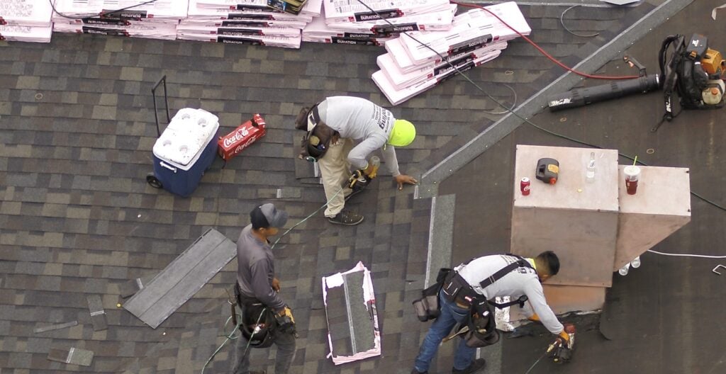 Three construction workers in safety gear tackle a roof shingle replacement