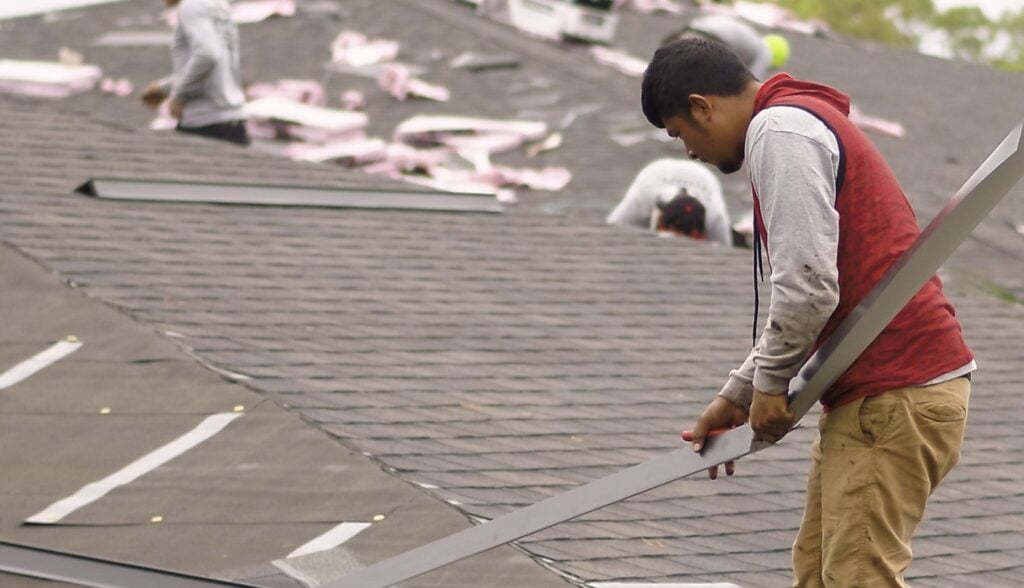 A worker in a red hoodie and tan pants installs shingles on a pitched roof while holding a long strip of material; another worker is seen on the roof in the background with roofing materials scattered around.