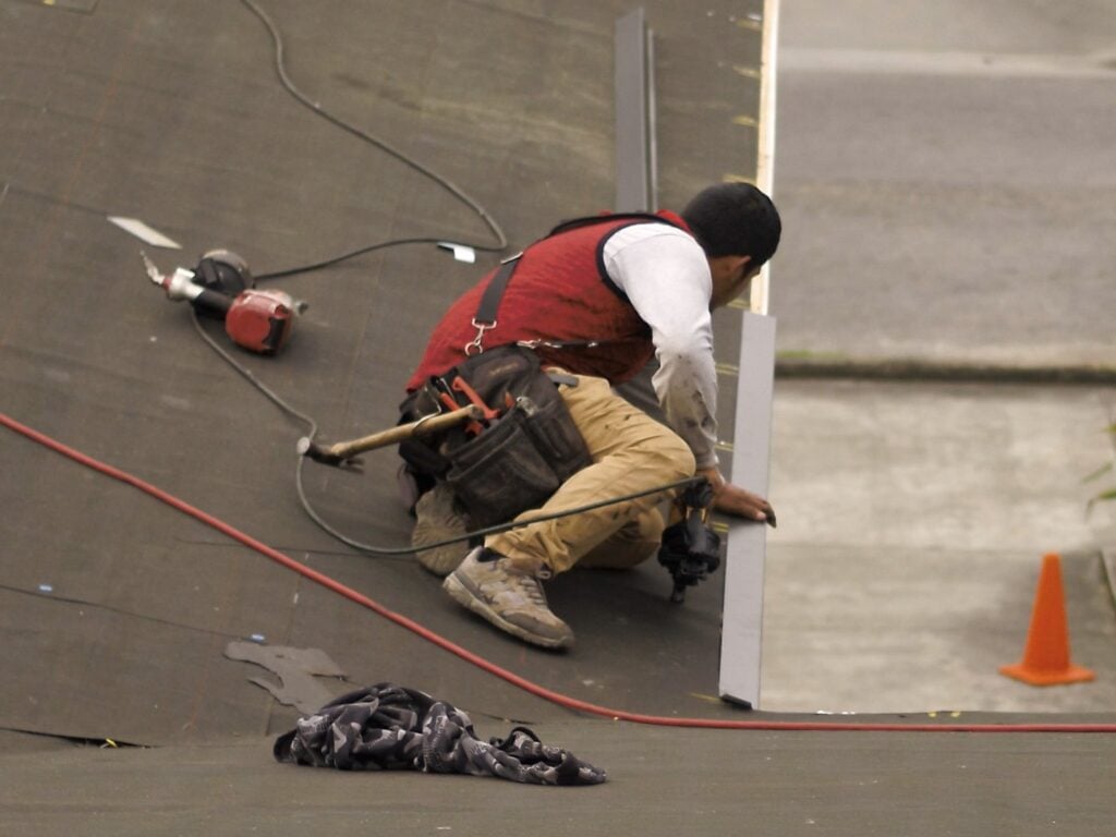 A construction worker wearing a red vest and tool belt kneels on a roof, measuring or installing material for a roof repair. Tools and a coiled cable are nearby, with a safety cone in the background, suggesting possible roof damage.