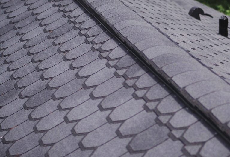 Close-up view of a gray shingle roof, showing overlapping asphalt shingles in neat rows along the ridge line. The surface is textured, and two roof vents are visible in the background to help improve airflow.