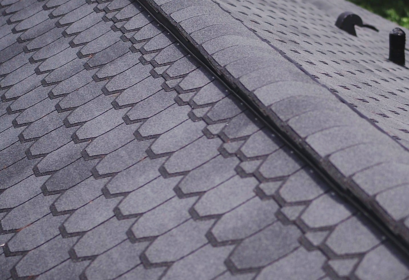 Close-up view of a gray shingle roof, showing overlapping asphalt shingles in neat rows along the ridge line. The surface is textured, and two roof vents are visible in the background to help improve airflow.