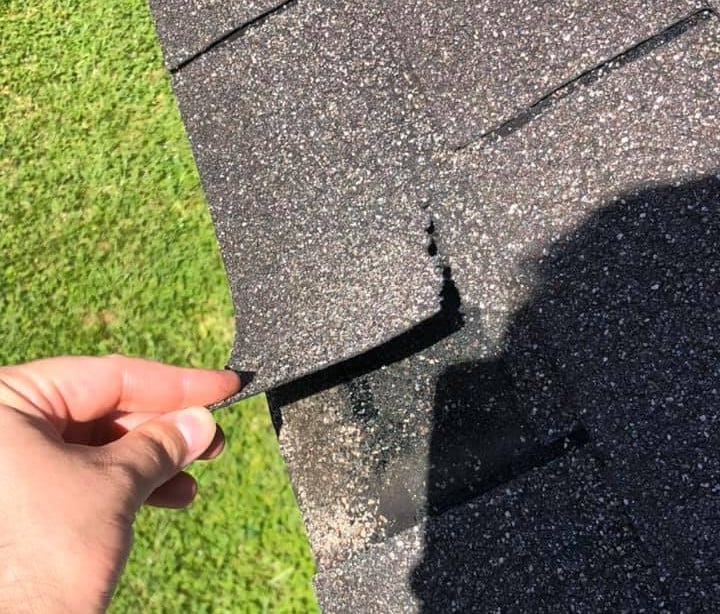 A hand lifts a loose asphalt shingle on a roof, revealing underlying roof damage