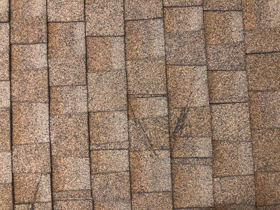 A close-up view of brown asphalt roof shingles arranged in overlapping rows, with a textured and slightly weathered appearance.