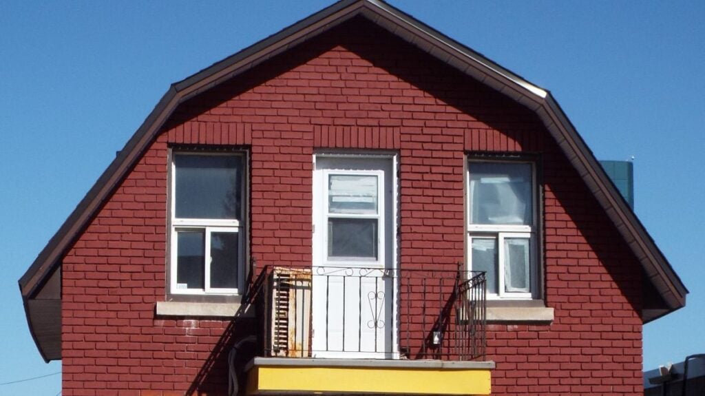 A red brick house with a stylish gambrel roof design, two windows, and a white door opening onto a small balcony with a metal railing, set against a clear blue sky.