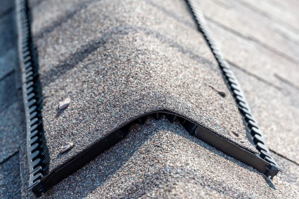 Close-up of a house roof ridge with asphalt shingles and a black ridge vent installed, highlighting the texture of the shingles and how roof vents at the peak enhance attic airflow for improved ventilation.