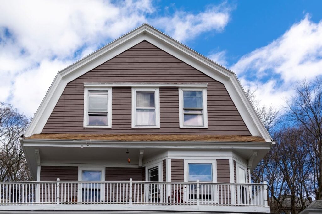 A two-story house with brown siding, white trim, and a wide front porch with a white railing features a classic gambrel roof design, set against a blue sky with scattered clouds and leafless trees—one of today's popular roof styles.