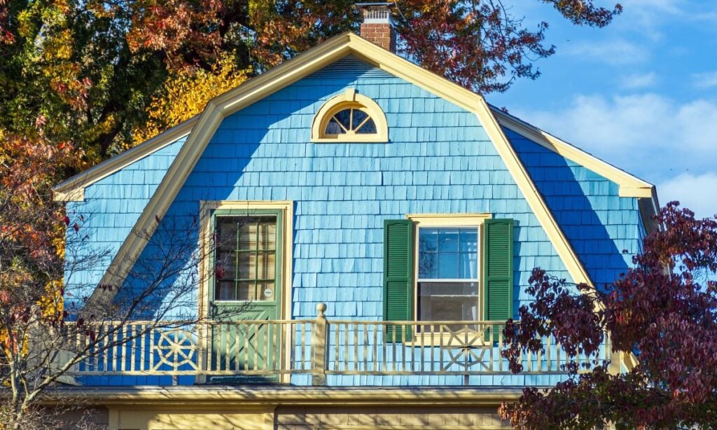 A close-up of a house with blue shingle siding, green shutters, and yellow trim. Notably, its gambrel roof complements the decorative upper window. Autumn trees with colorful leaves surround the home under a partly cloudy sky.