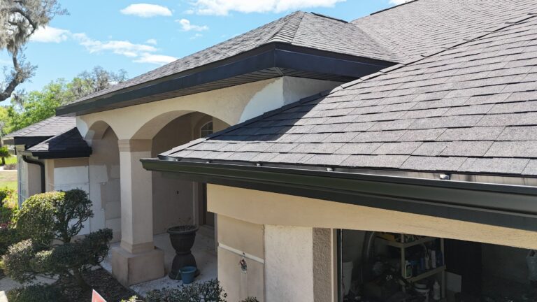 A close-up view of a house exterior shows dark-colored shingles on the roof, black gutters and fascia, beige stucco walls, and landscaped bushes along the walkway under a partly cloudy sky.