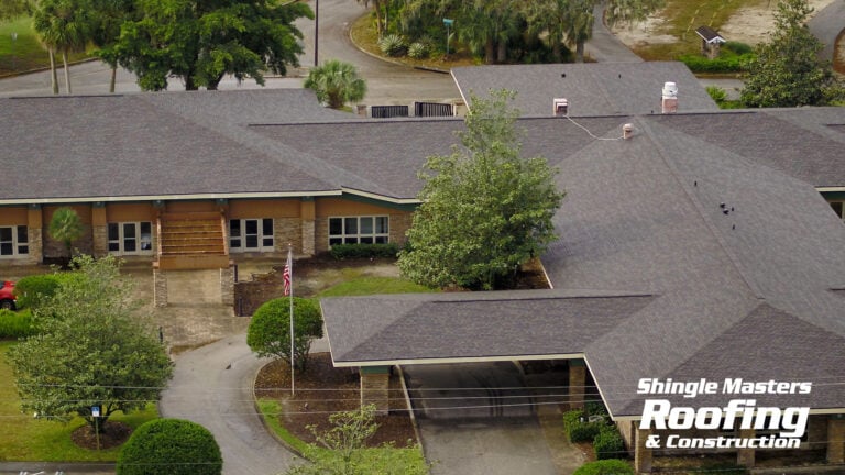 Aerial view of a large building with a dark shingle roof, ideal for commercial roofing or multi family roofing projects, surrounded by trees and landscaping. The logo "Shingle Masters Roofing & Construction" appears in the bottom right corner.
