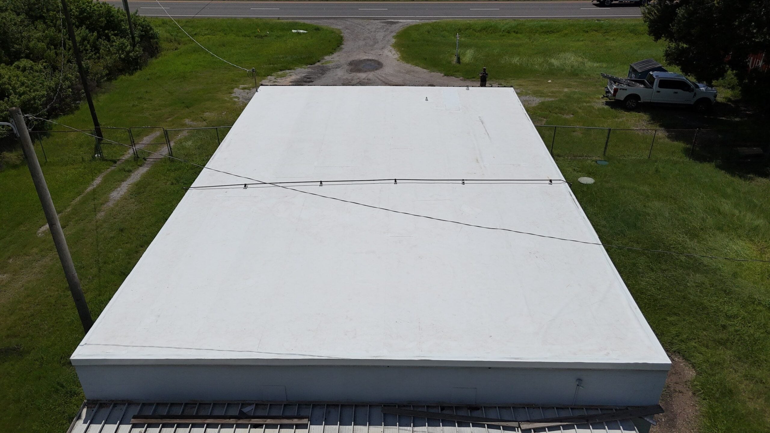 Aerial view of a flat white-roofed building, ideal for commercial roofing, surrounded by grass, with a parked white truck and a road in the background. Power lines cross over the roof.