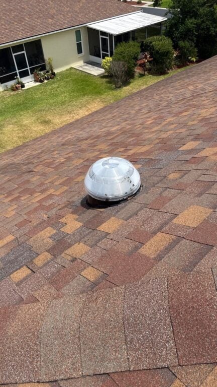 A dome-shaped skylight is installed on a brown shingled roof with well-maintained fascia, while a grassy yard and neighboring houses are visible in the background.