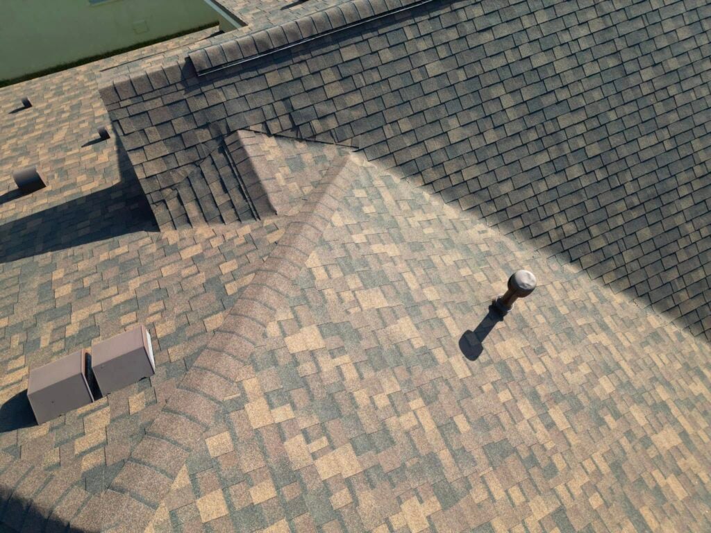 Aerial view of a house roof covered with patterned composite asphalt shingles in brown and green shades, showcasing ridges, a vent pipe, and two roof vents—details homeowners should note to avoid common mistakes on roof insurance claims.