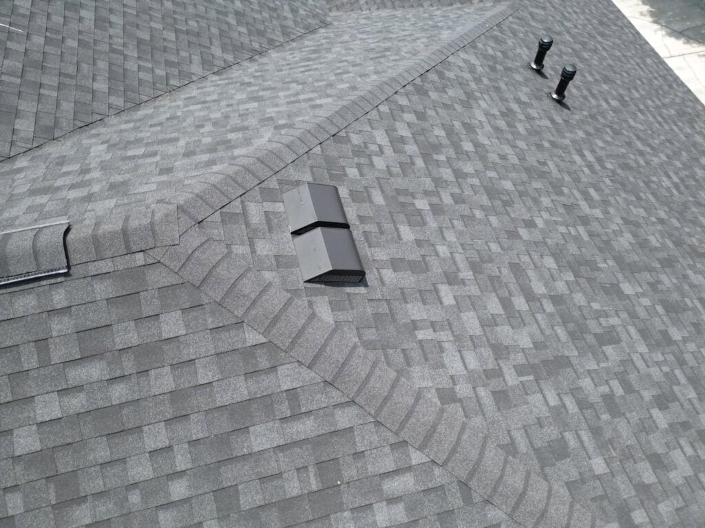 A close-up view of a gray asphalt shingle roof featuring a Slant Back Roof Vent, ridge vent, and intersecting rooflines with clean, uniform shingle patterns that highlight the performance benefits of proper ventilation.