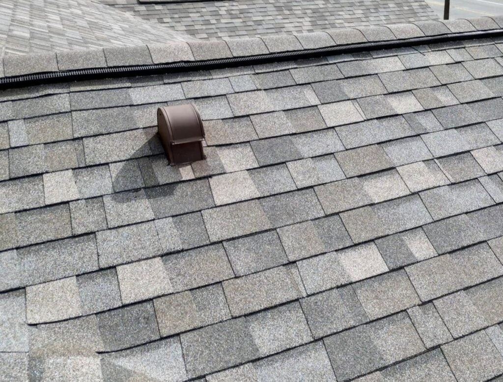 Close-up view of a shingled roof in gray and brown shades featuring a Slant Back Roof Vent near the ridge, highlighting its performance benefits. Portions of neighboring roof sections are visible in the background.