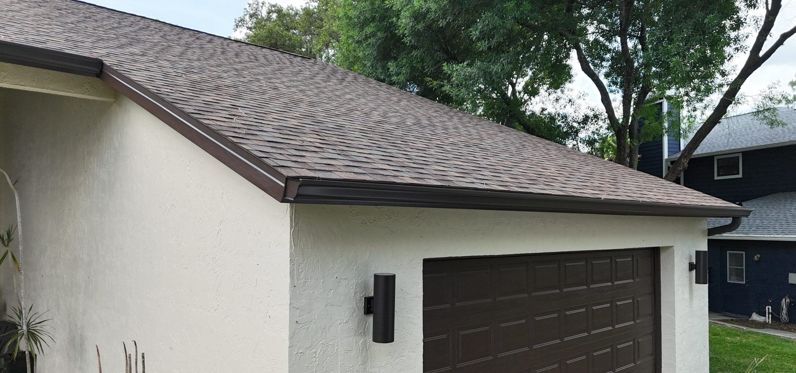 A cream-colored garage with a dark brown door and matching trim, including sleek gutters and fascia, features two modern black wall lights. Trees and part of a neighboring house are visible in the background.