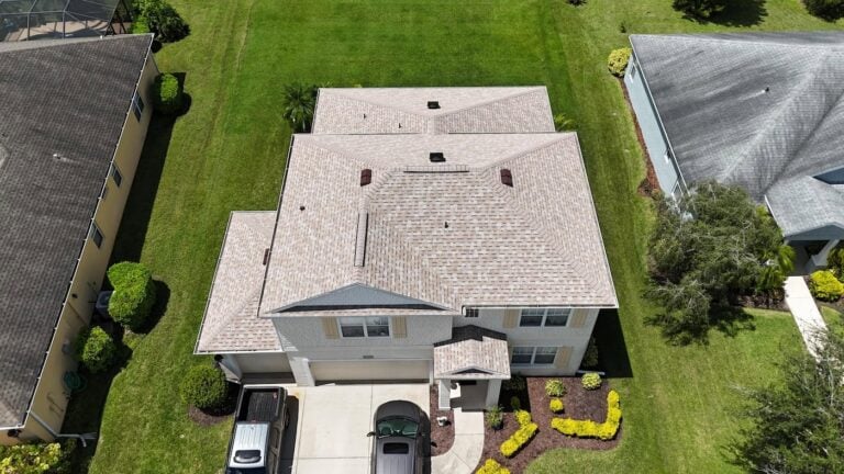 Aerial view of a two-story house with a light-colored roof, driveway with two cars parked, well-maintained lawn, and landscaped shrubs. The house is situated between two similar homes in a suburban neighborhood.