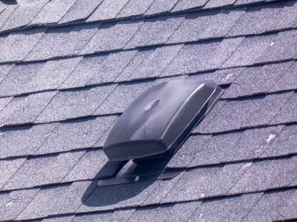 A close-up of a rooftop with gray asphalt shingles and a black plastic slant back roof vent installed near the center, highlighting both the installation process and performance benefits.