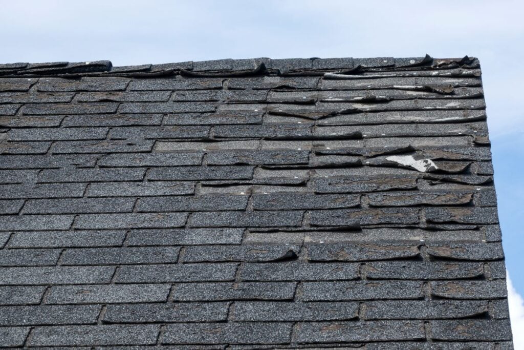 A roof with dark asphalt shingles, some curled, bent, and damaged—roof warning signs that may point to weather damage or causes of granular loss—sits under a partly cloudy sky.