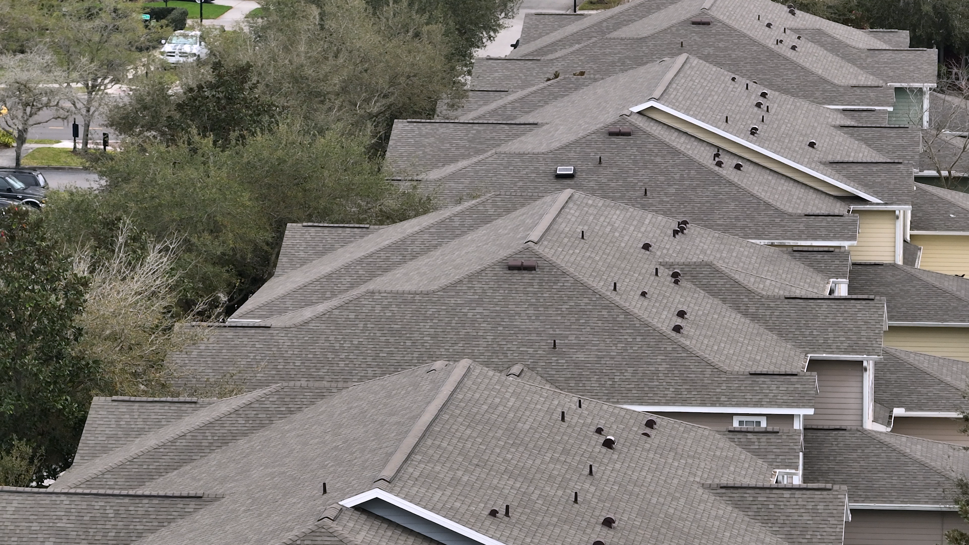 Row of closely packed suburban houses with gray shingled roofs, seen from above. Trees border the houses, and a street is visible in the background. The multi family roofing creates a repetitive, geometric pattern.