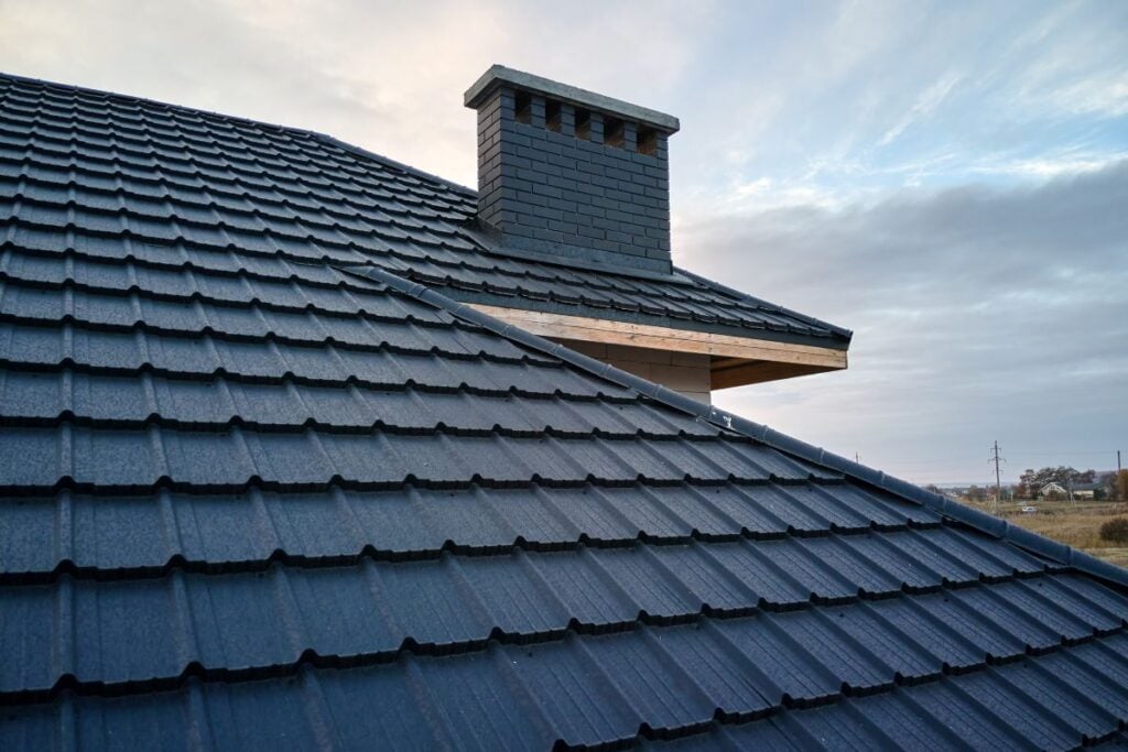 Close-up view of a dark blue tiled roof with a brick chimney under a cloudy sky. This scene highlights the importance of choosing the right option, as showcased in any homeowner’s guide to different metal roof types and styles.
