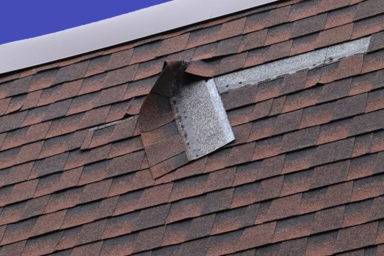 Close-up of a roof with reddish-brown asphalt shingles. Exposed, bent flashing suggests storm or wind damage—an ideal scenario for those seeking a roof repair guide or tips on repairing roof without replacement, against a clear blue sky.