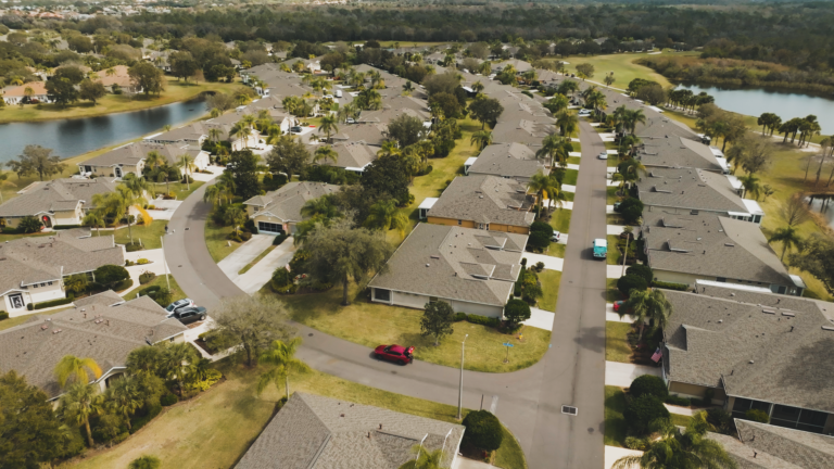 Aerial view of a suburban neighborhood with curved streets, single-story houses featuring well-maintained fascia and gutters, well-kept lawns, palm trees, and cars in driveways. A lake and open green spaces appear in the background.