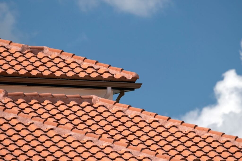 Close-up view of a red clay tiled roof against a blue sky with a few white clouds. The neatly arranged tiles highlight tile roof durability, covering multiple roof sections with lasting protection.