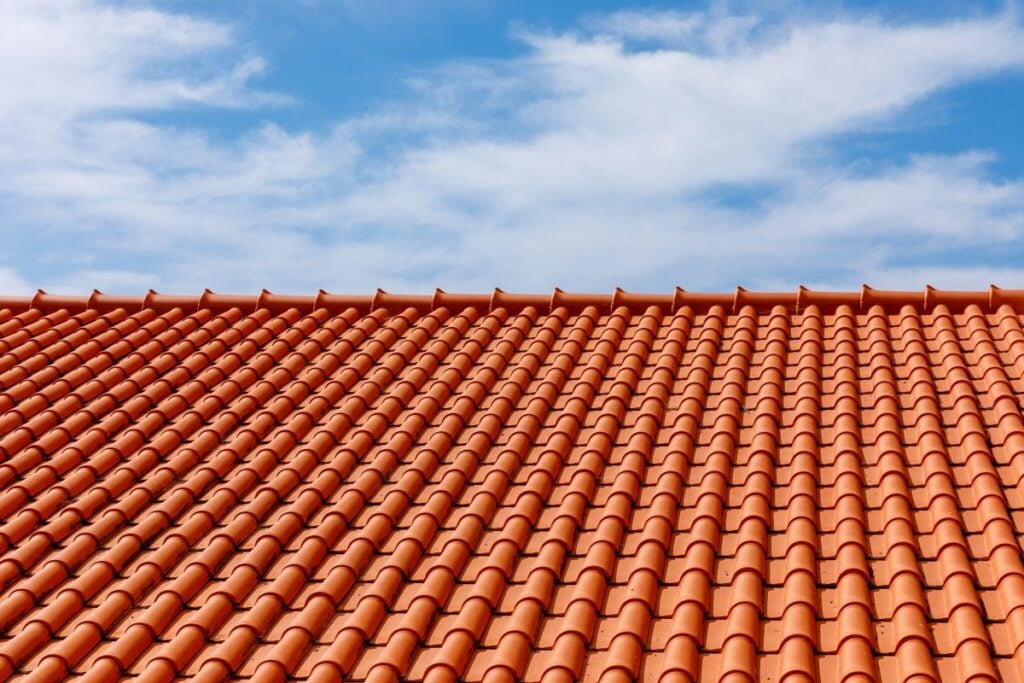 A close-up view of an orange clay tile roof under a blue sky with scattered white clouds. The neatly arranged rows highlight tile roof durability and the orderly pattern that contributes to the impressive tile roof lifespan.