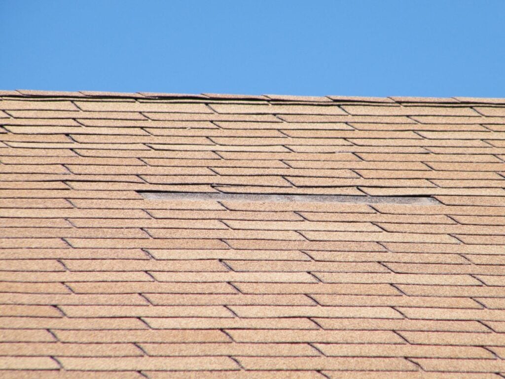 A close-up of a shingled roof under a clear blue sky, highlighting an area with missing or damaged shingles—an ideal example for a roof repair guide on how to repair damaged roof sections without replacing the entire surface.