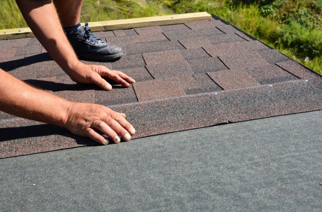 A person repairs a damaged roof by installing asphalt shingles, carefully aligning them by hand. Black underlayment covers part of the roof, while green grass can be seen in the background—a practical step from any roof repair guide.