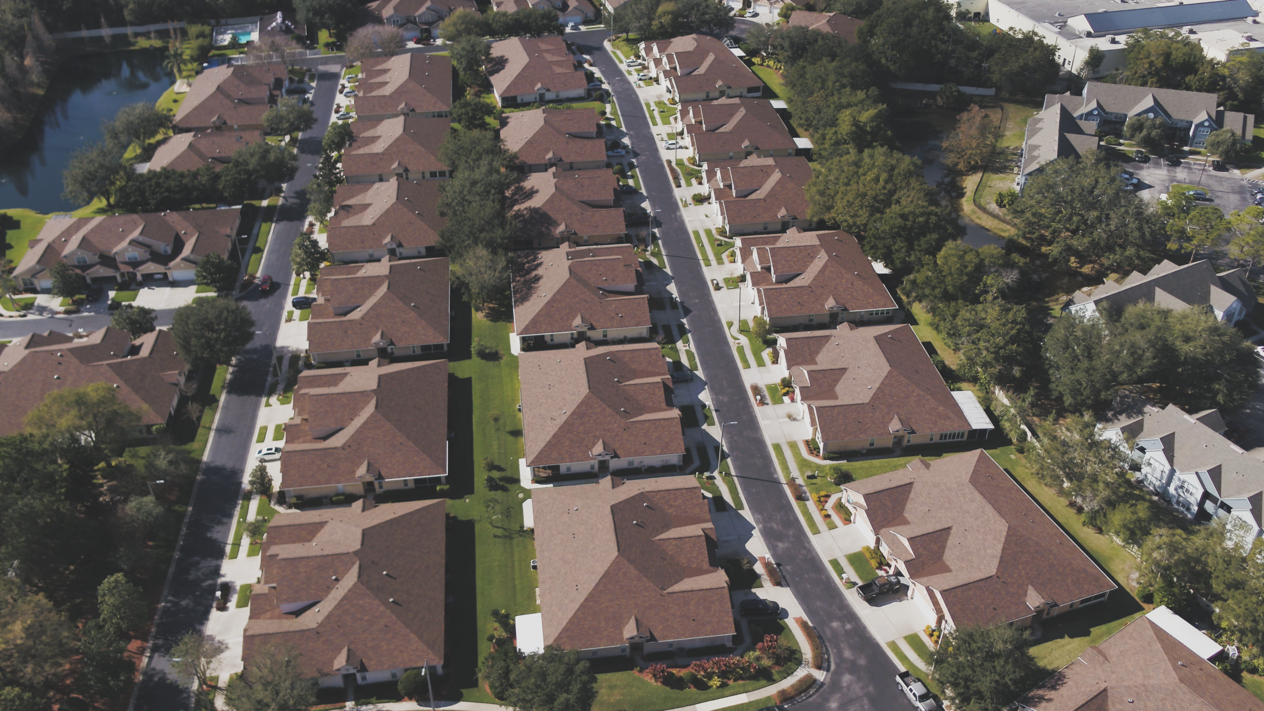 Aerial view of a suburban neighborhood with rows of identical houses featuring brown roofs, driveways, green lawns, and tree-lined streets on a clear day—highlighting uniformity ideal for multi family roofing solutions.