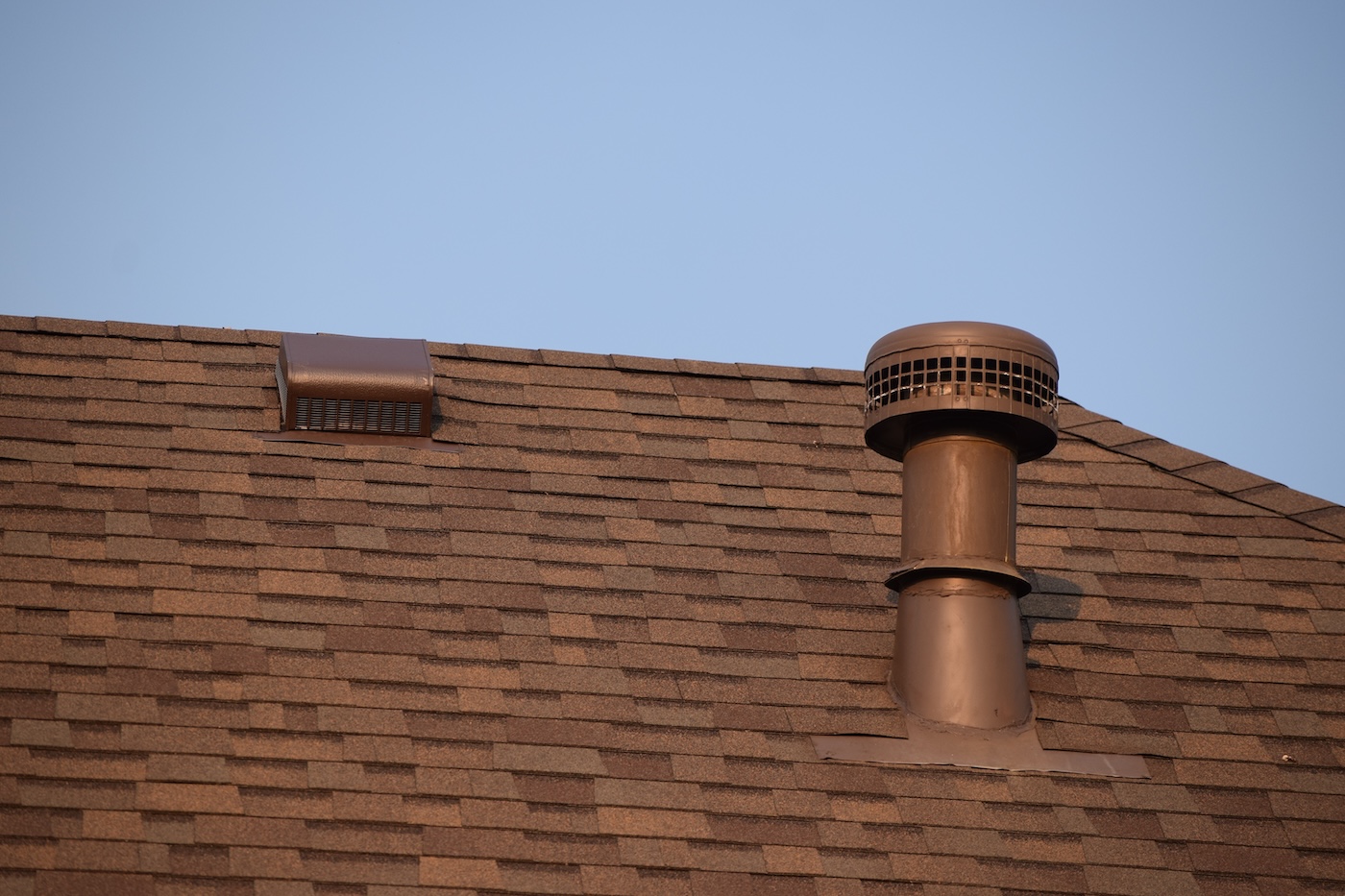 A close-up of a brown shingled roof with a metal vent pipe and a rectangular vent cover, set against a clear blue sky.