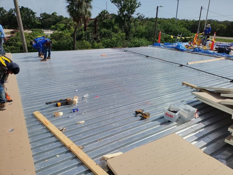 Workers on a metal rooftop installing materials for a multi family roofing project, with tools, wood planks, and insulation sheets spread out; trees and power lines are visible in the background.