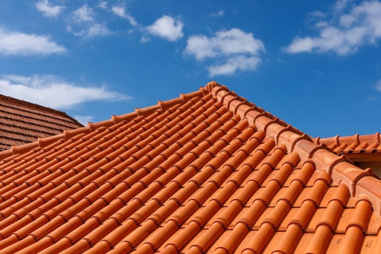 Close-up view of a house roof with orange clay tiles against a blue sky with a few white clouds. The tile roofing is neatly arranged in rows, creating a textured, uniform pattern.