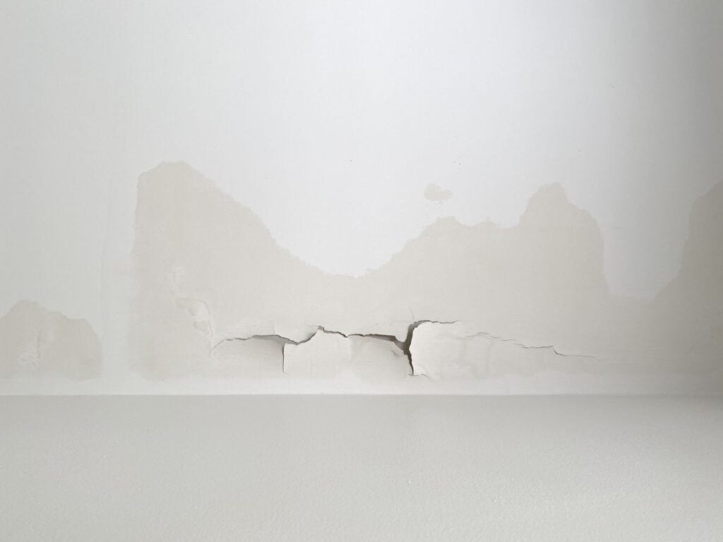 A white wall with large areas of peeling and cracked paint, showing signs of water damage and discoloration—points that could indicate the need for roof leak repair or tips to prevent roof damage. The damaged section has multiple cracks and uneven surfaces.
