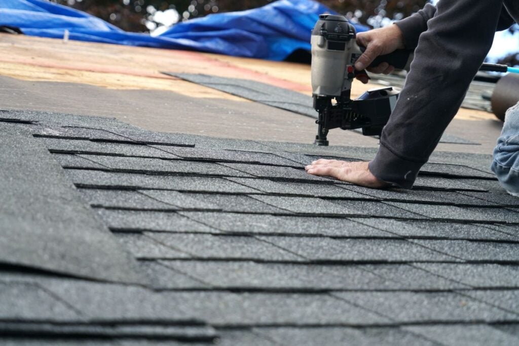 A person installs asphalt shingles on a roof using a nail gun, pressing down on the shingles for a secure fit, helping with roof leak prevention. The roof and nearby blue tarp are visible.