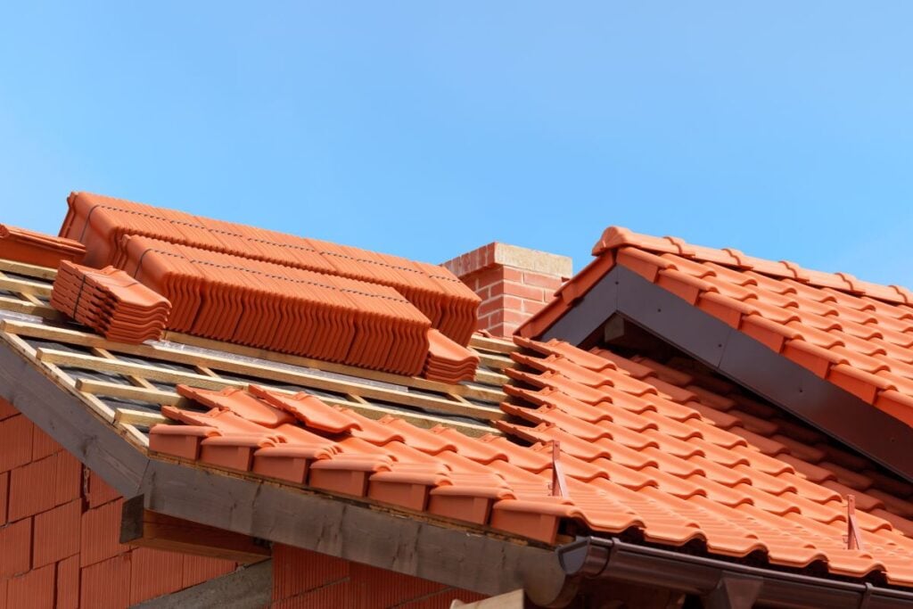 Stacks of red clay roof tiles are placed on the wooden framework of a partially constructed roof, demonstrating effective tile roof tips. A brick chimney and clear blue sky are visible in the background.