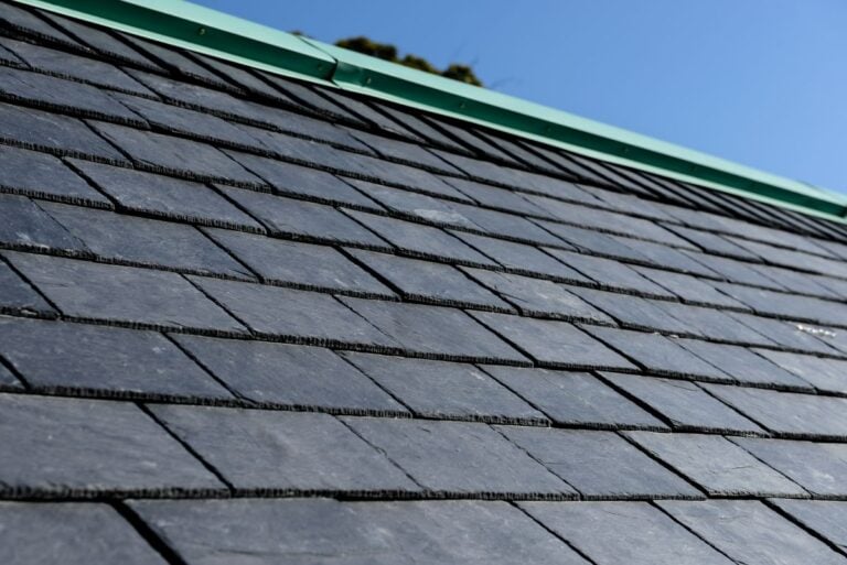 Close-up view of a dark gray slate tile roof with a green metal ridge cap, set against a clear blue sky—highlighting the impressive tile roof durability.