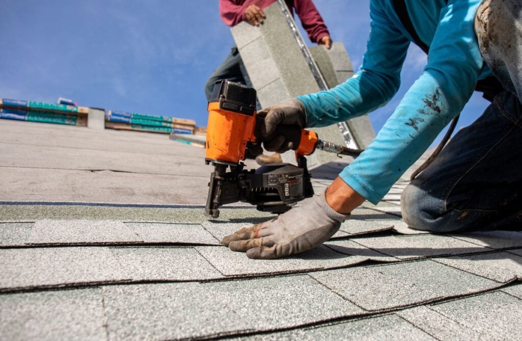 A close-up of a worker using a nail gun to install asphalt shingles, with another worker holding shingles in the background. Both wear gloves and long sleeves under a clear blue sky, illustrating steps from a roof repair guide to fix roof without replacement.