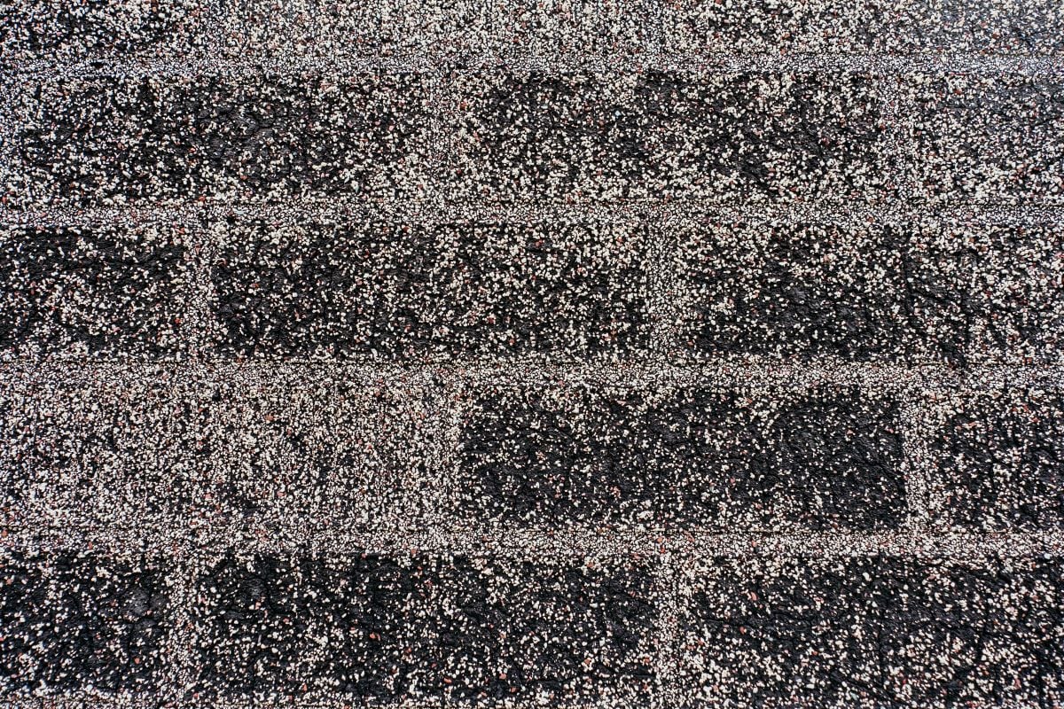 Close-up of dark asphalt roof shingles with a textured surface, featuring horizontal rows and light-colored granules scattered across the surface—a common sign of granular loss on roof and one of the early roof warning signs.