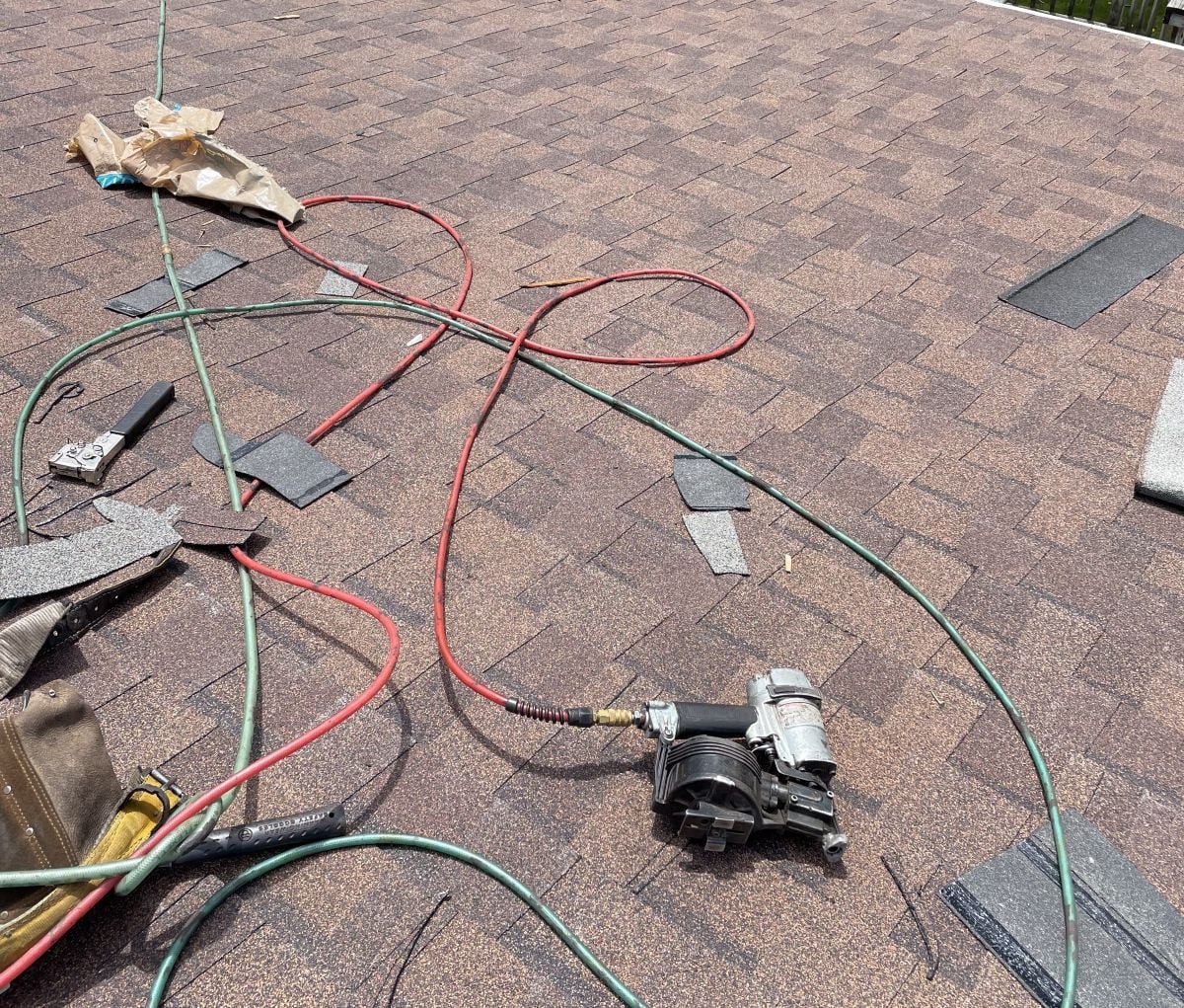 A roofing nail gun with attached hoses lies on a brown shingle roof, surrounded by scattered shingles, a tool pouch, and a paper bag—evidence of ongoing roof leak repair to prevent roof damage.