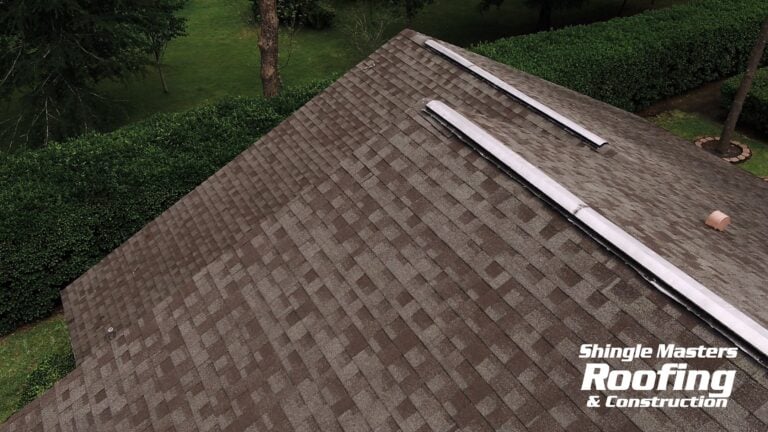 Aerial view of a brown shingle roof with two vent pipes, showcasing the best shingle color choices for a Florida home, surrounded by green trees and bushes. "Shingle Masters Roofing & Construction" logo appears in the lower right corner.