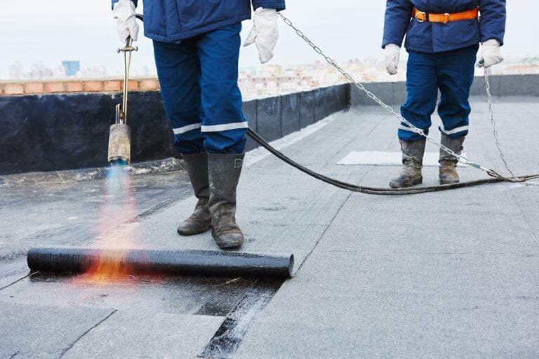 Two workers in protective gear use a propane torch to install a waterproof membrane on a flat rooftop, applying heat to roll out and seal the material