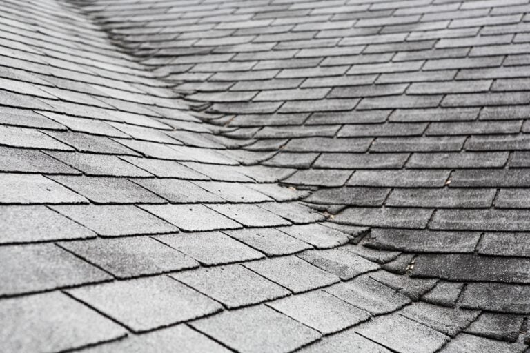 Close-up of an asphalt shingle roof showing visible sagging and unevenness, indicating possible structural issues. The gray shingles appear weathered, with some shingles curling—a common sign that early fixes for shingles may be needed to prevent further damage.