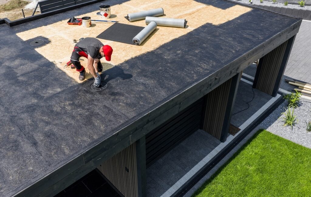 A construction worker is installing EPDM roofing material on a contemporary home under clear skies, surrounded by tools and prepared materials for the roofing project.