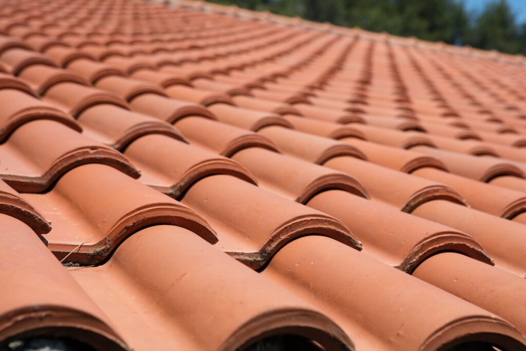 Close-up of a red clay tile roof, with curved tiles arranged in overlapping rows, creating a repeating pattern typical of traditional clay tile roofs. The background is blurred with some green trees visible.